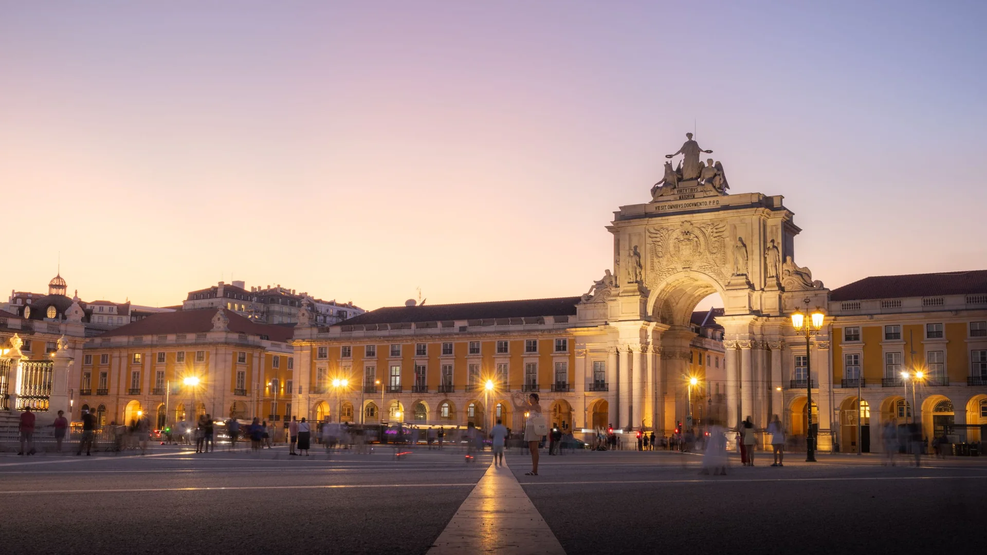 Praça do Comércio's yellow arcades and the Rua Augusta Arch lit at twilight, with a few people crossing the empty square
