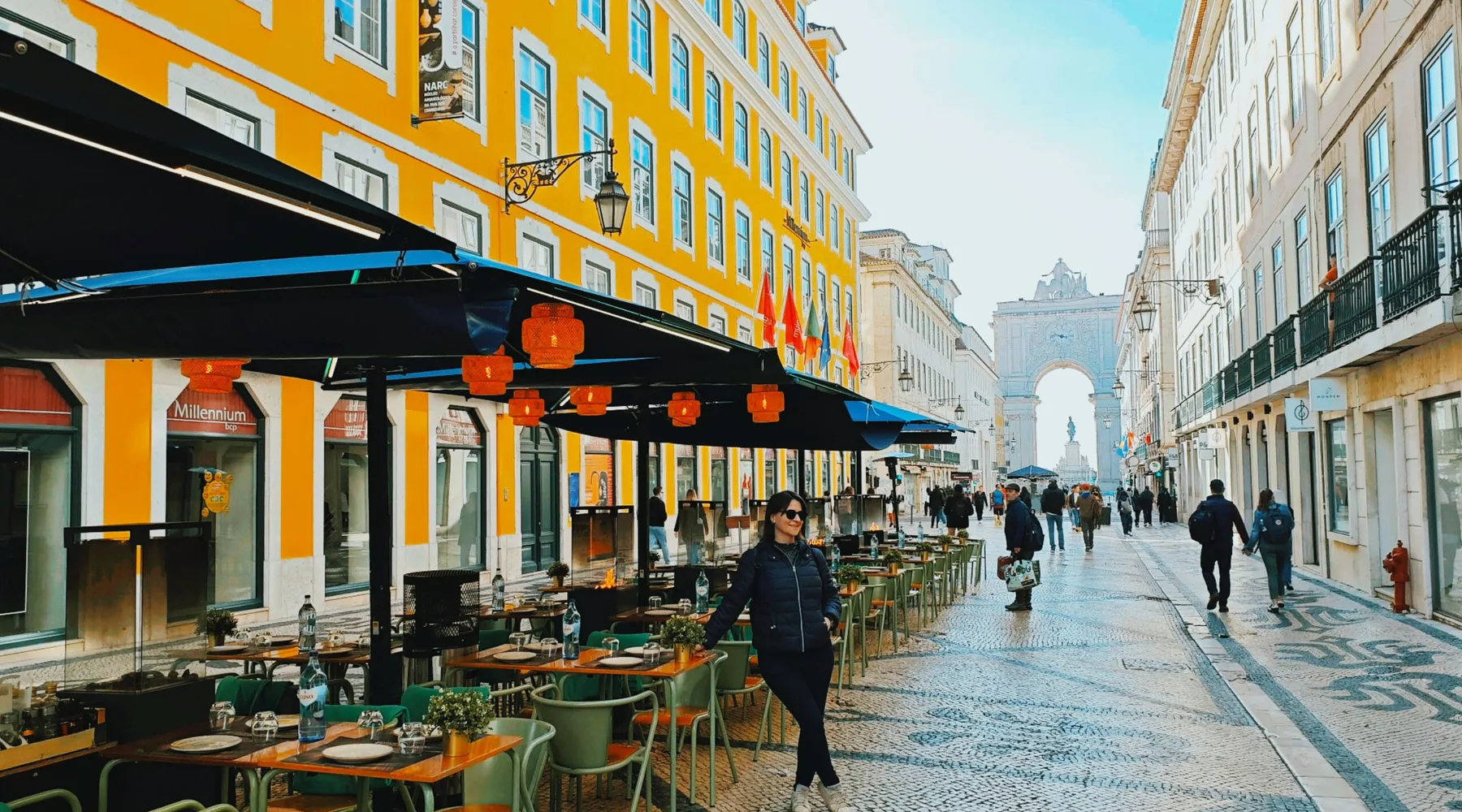 Rua Augusta in Lisbon's Baixa, café terraces under yellow façades on the left and the Rua Augusta Arch at the far end of the cobbled street